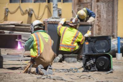 Two construction workers in safety vests and hard hats operating welding equipment on a job site.