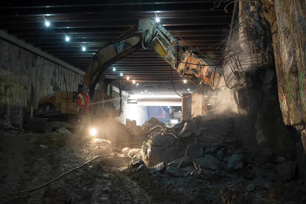Excavator with jackhammer attachment removing concrete in underground station construction area with dust and work lights.