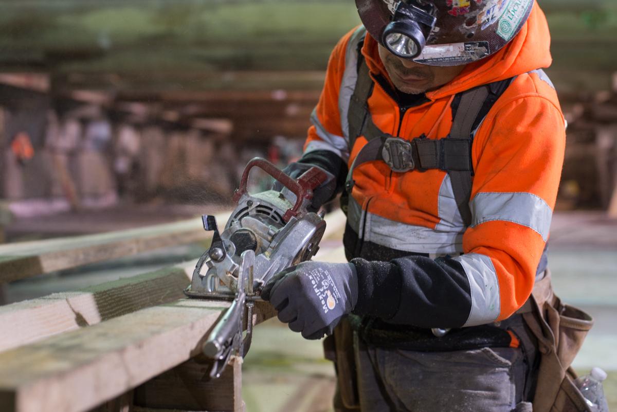Worker in orange safety vest operating a power saw on wooden construction materials at station platform level.