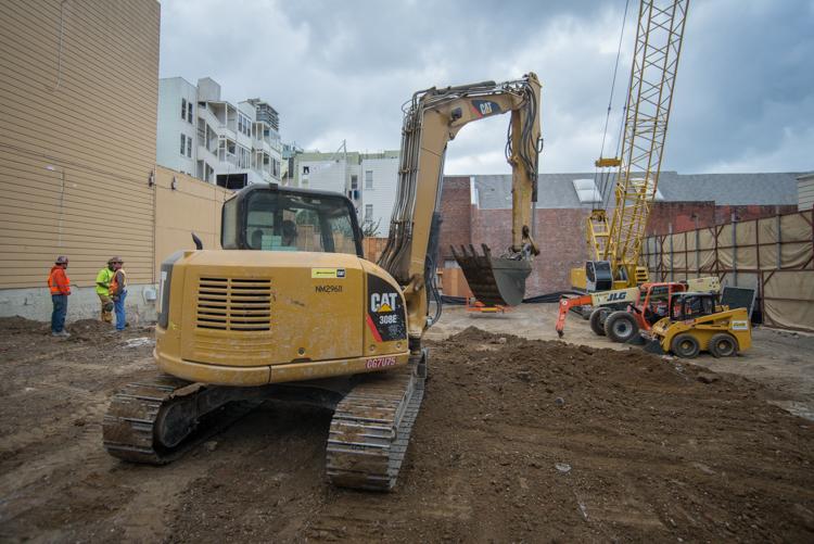 Yellow CAT excavator on construction site with workers, crane, and buildings in background.