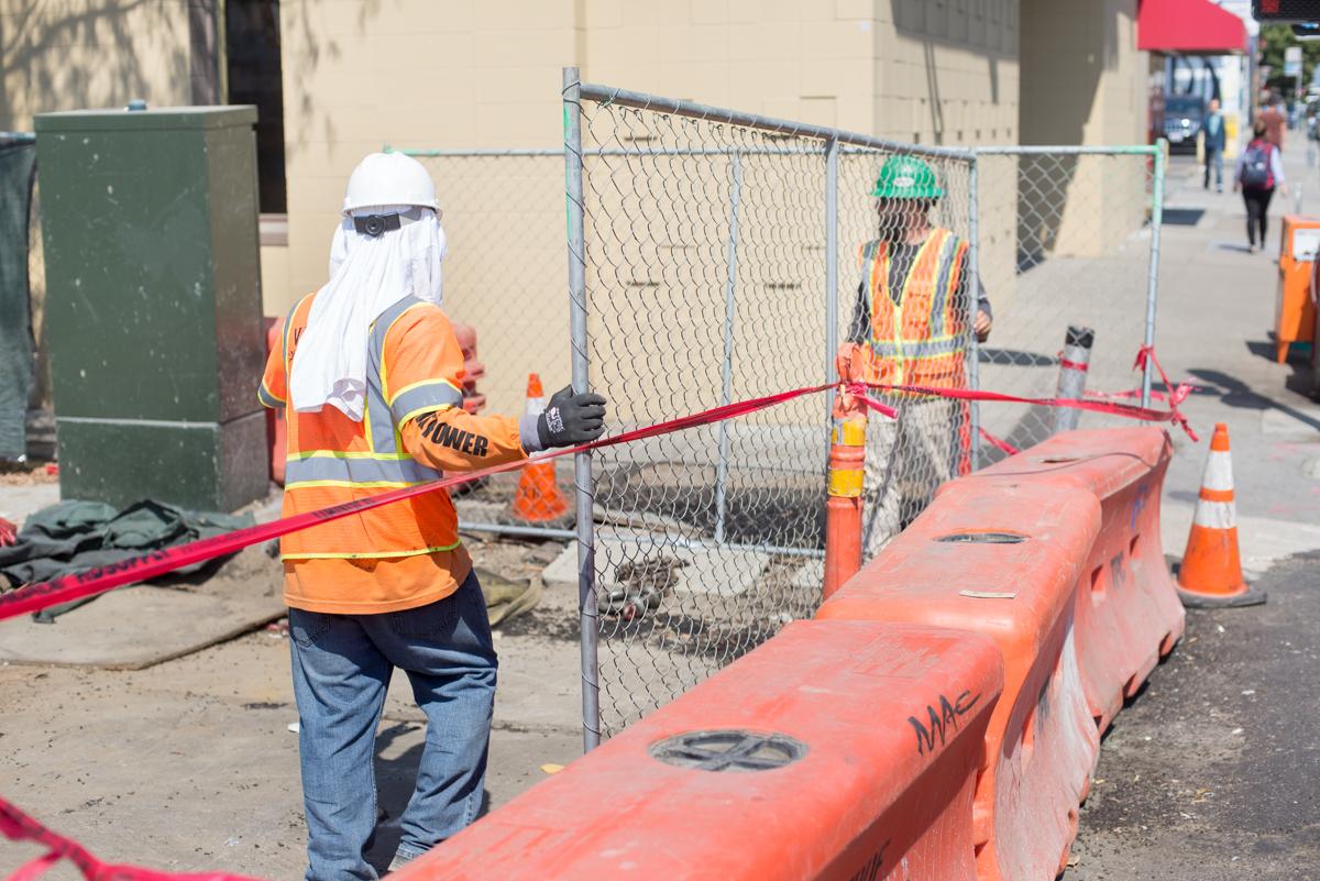 Construction workers in safety vests set up a chain-link fence and red barriers at a street work site.