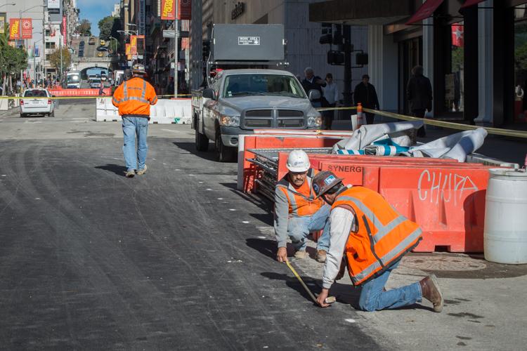 Construction workers in orange safety vests and hard hats work on Stockton Street with traffic barriers and equipment visible.