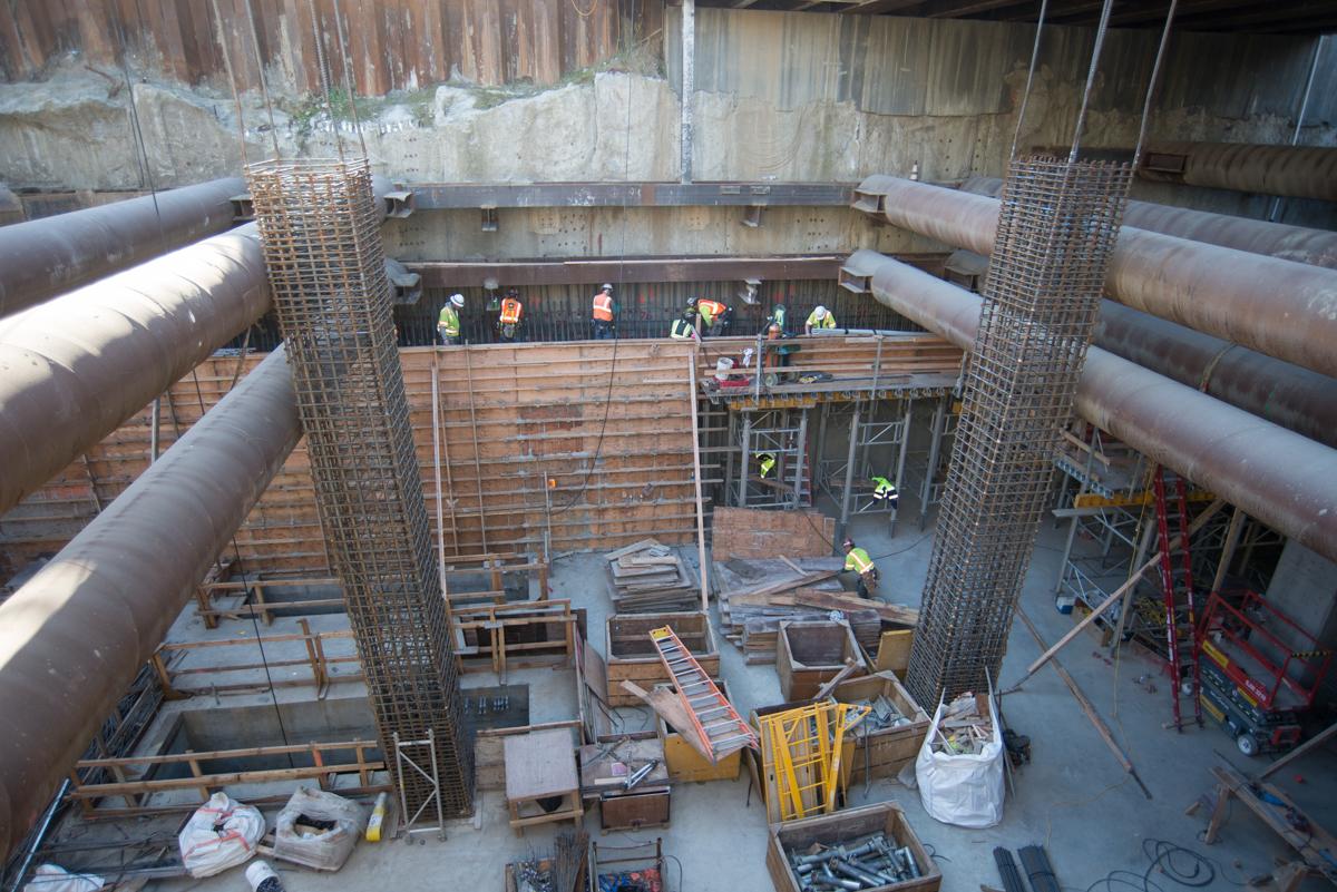 Construction site showing reinforced concrete column cages and workers inside underground transit station headhouse.