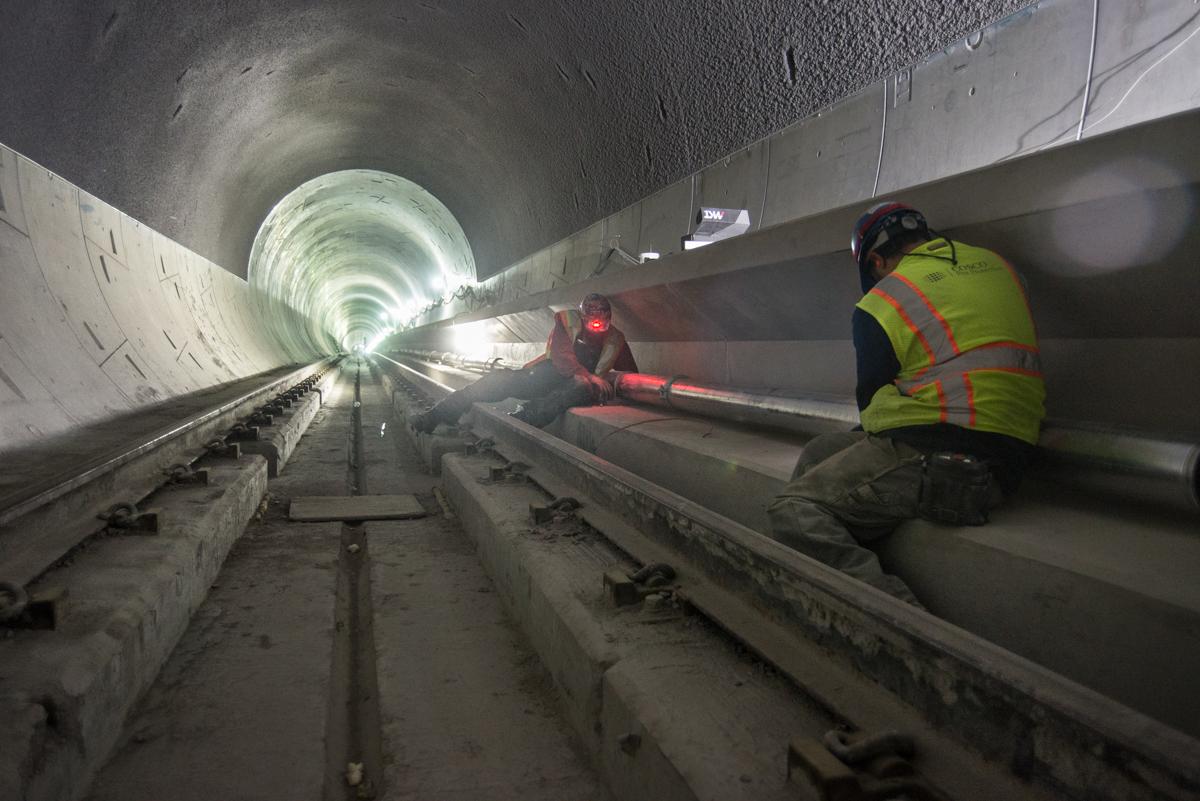 Workers in safety vests install utility conduit along emergency walkway inside subway tunnel under construction.