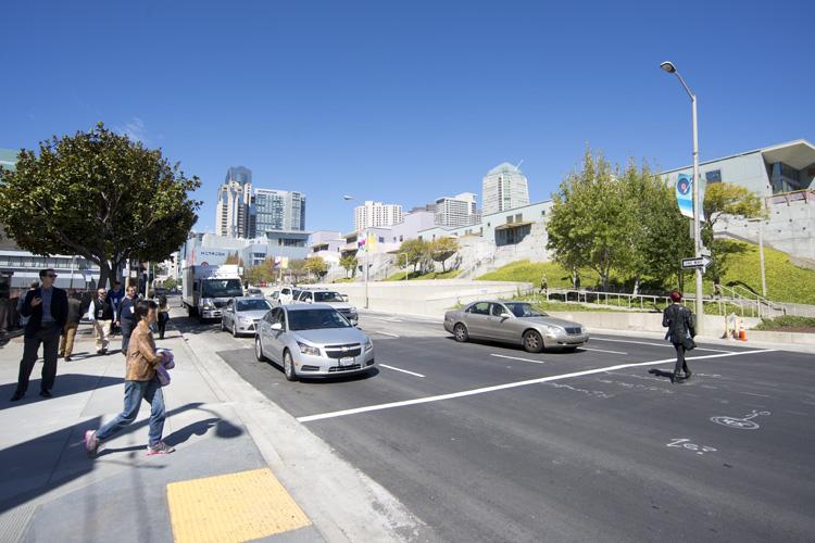 Busy urban street intersection with pedestrians crossing, parked and moving vehicles, trees, street lamp, and downtown skyline in background under clear blue sky.