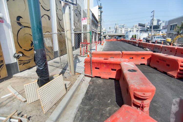Construction site with orange barriers and asphalt work on urban street with city skyline in background.