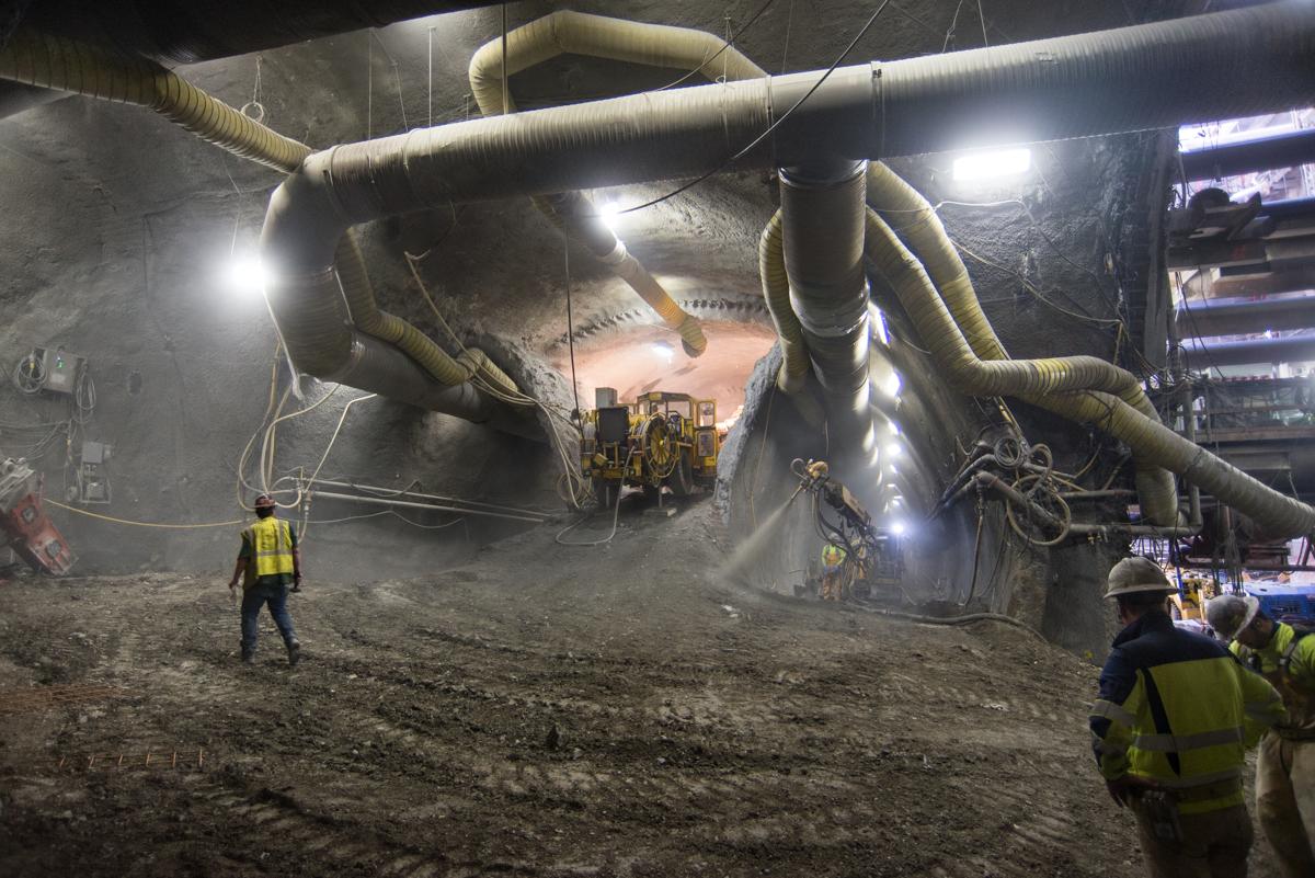 Workers in safety vests conduct underground tunnel construction with large pipes and heavy equipment in an illuminated cavern.