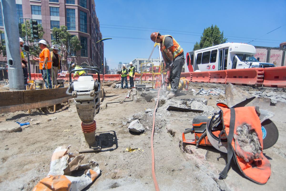 Construction workers jackhammer concrete around a manhole on 4th Street with equipment and barriers visible.