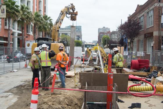 Construction workers in safety gear operate excavation equipment and install sheet piles during Central Subway trench work on an urban street.
