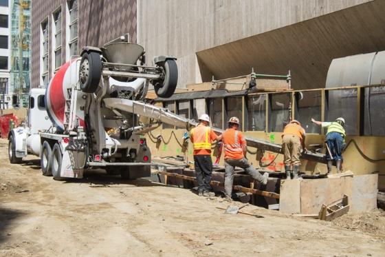 Construction workers pour cement from a concrete mixer truck at a transit station construction site in an urban area.
