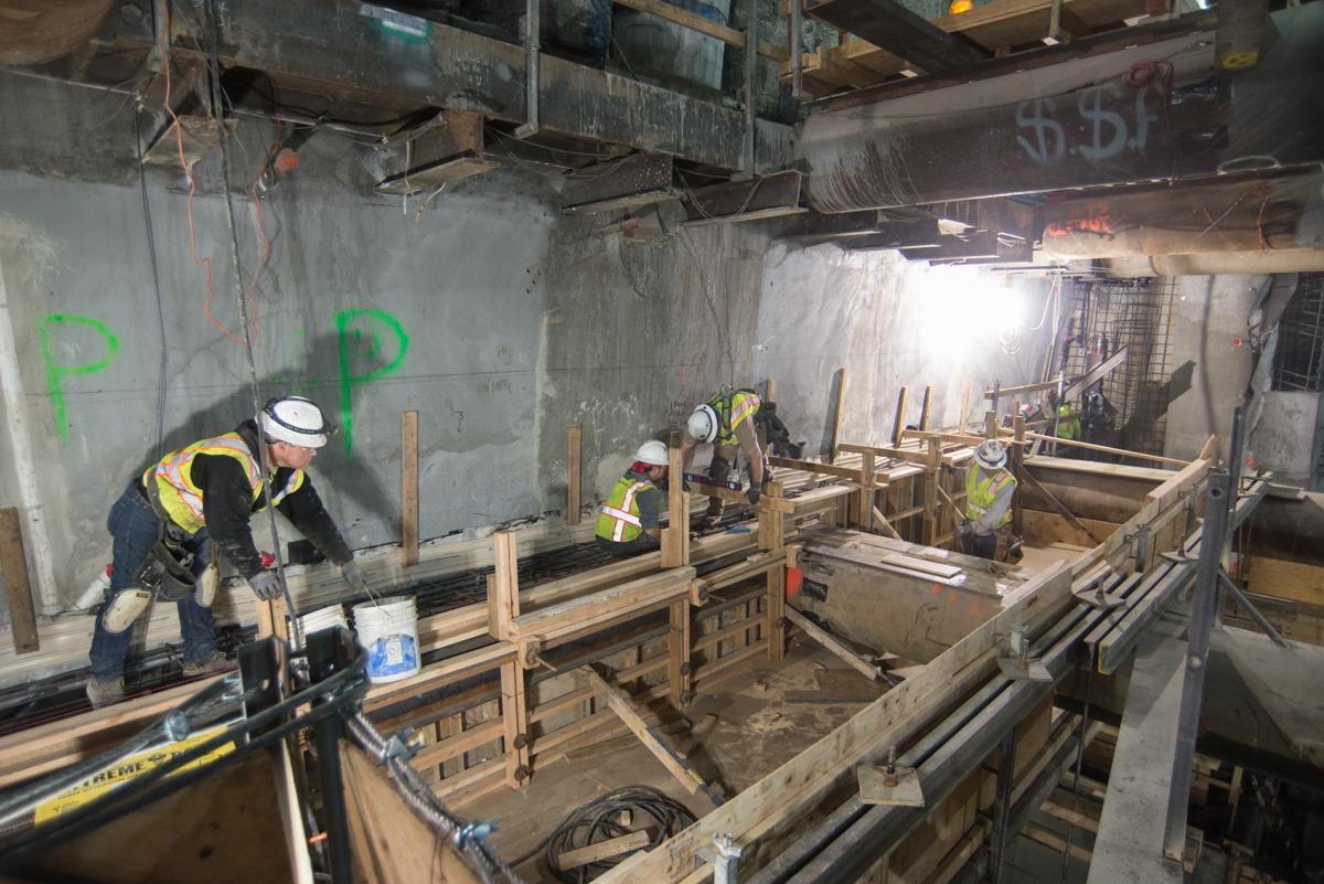 Construction workers in hard hats and safety vests build wooden concrete forms in an underground transit tunnel.