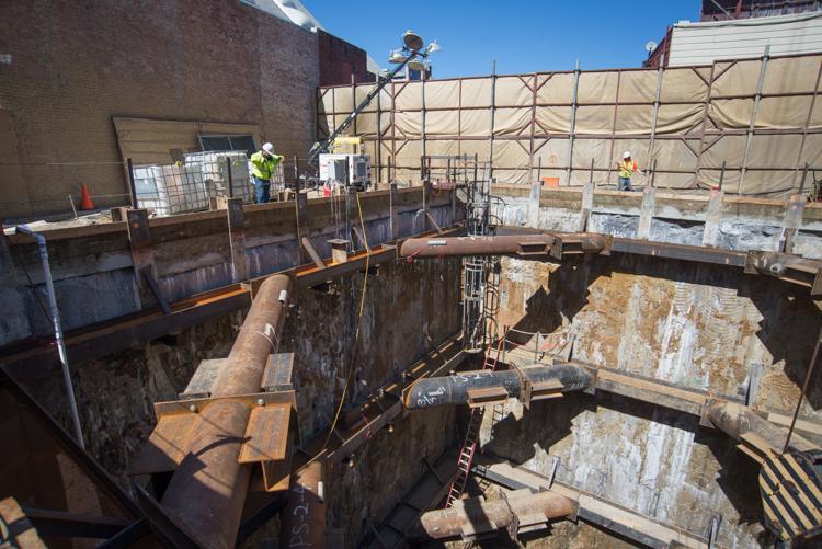 Construction site showing tunnel boring machine retrieval shaft with temporary cross-braces, metal framework, and workers in safety gear.