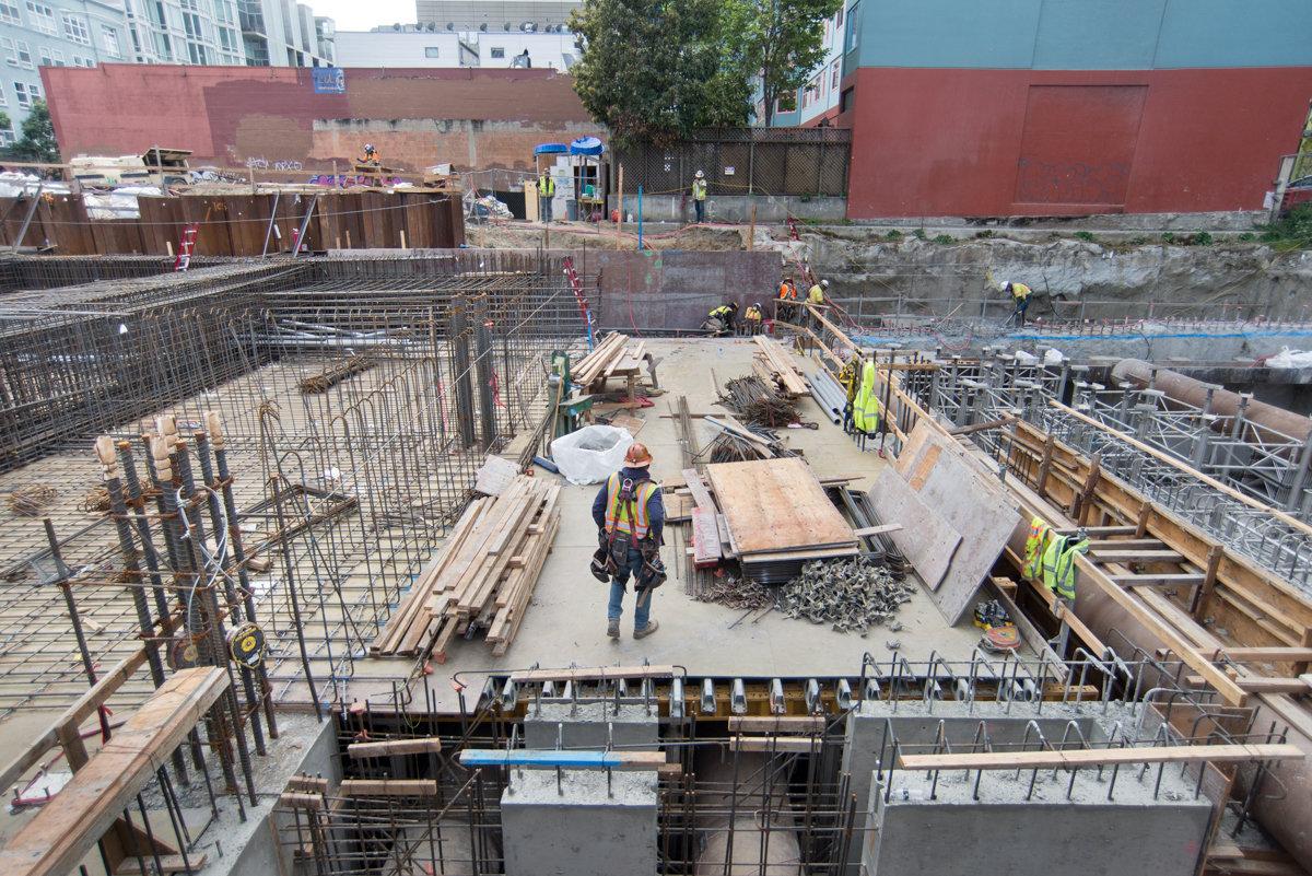Construction site showing excavation pit with reinforced concrete foundation, steel rebar framework, and workers in safety gear at Yerba Buena/Moscone Station.