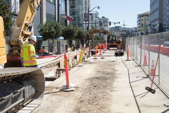 Construction site on urban street with excavator, safety barriers, and workers in hard hats.
