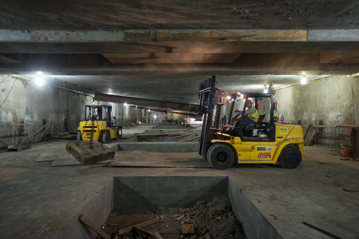 Yellow forklifts remove steel bracing from underground station construction site with concrete walls and overhead beams.