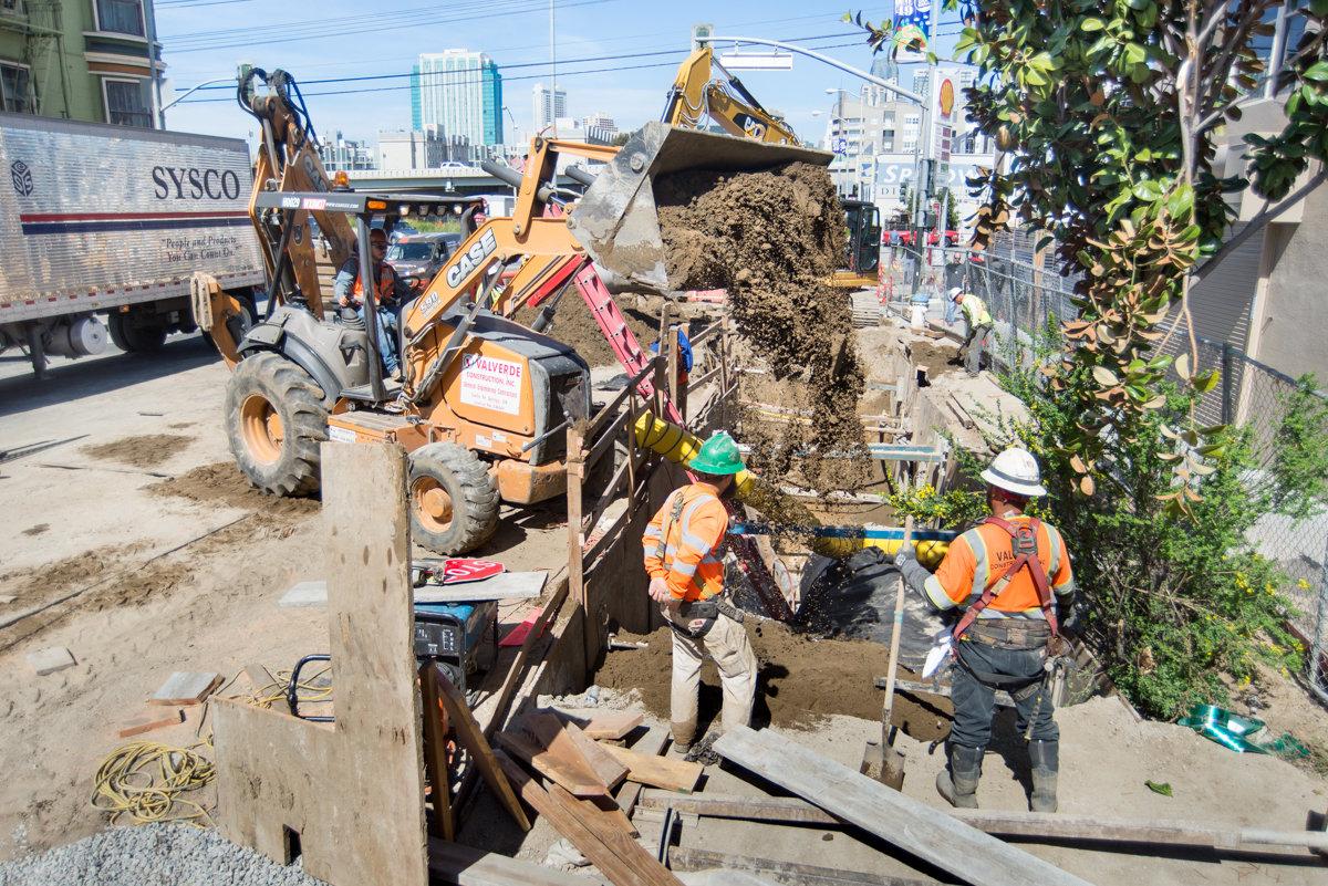 Construction workers and backhoe operator backfilling utility trench on urban street.