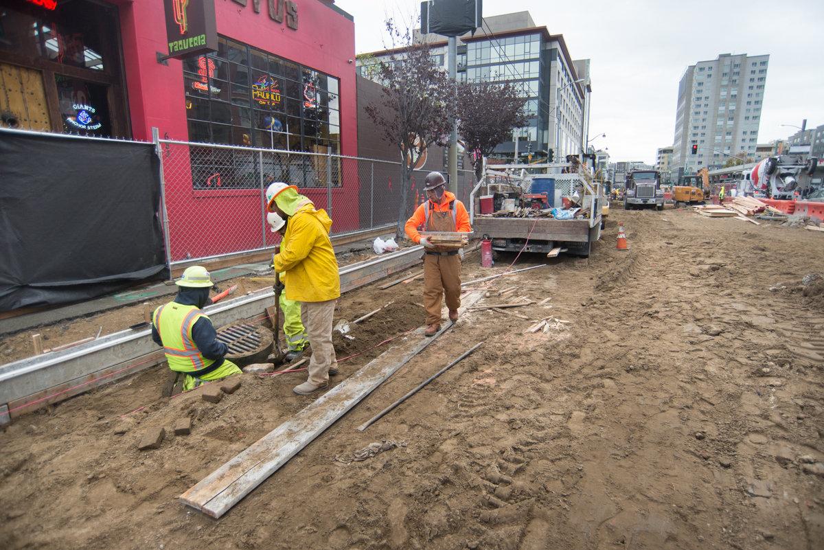Construction workers in safety vests install track and foundation work on a city street surrounded by buildings and equipment.