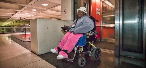 Person in wheelchair with pink blanket using elevator at underground transit station.