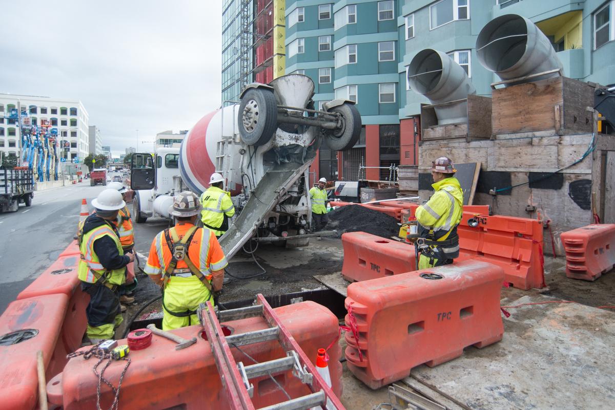 Construction workers in safety gear operate a concrete mixer truck at a transit station excavation site.
