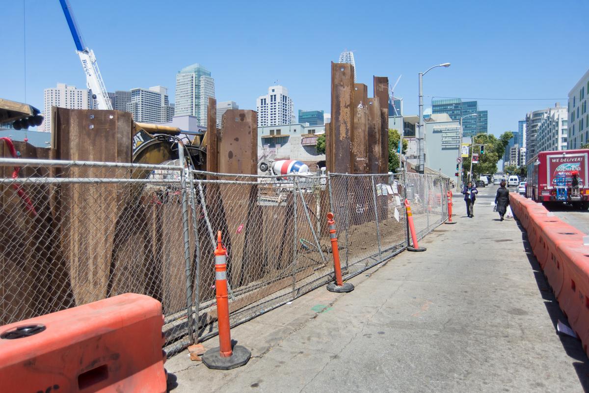Construction site at 4th and Folsom station with steel sheet piles, fencing, and downtown skyline visible.