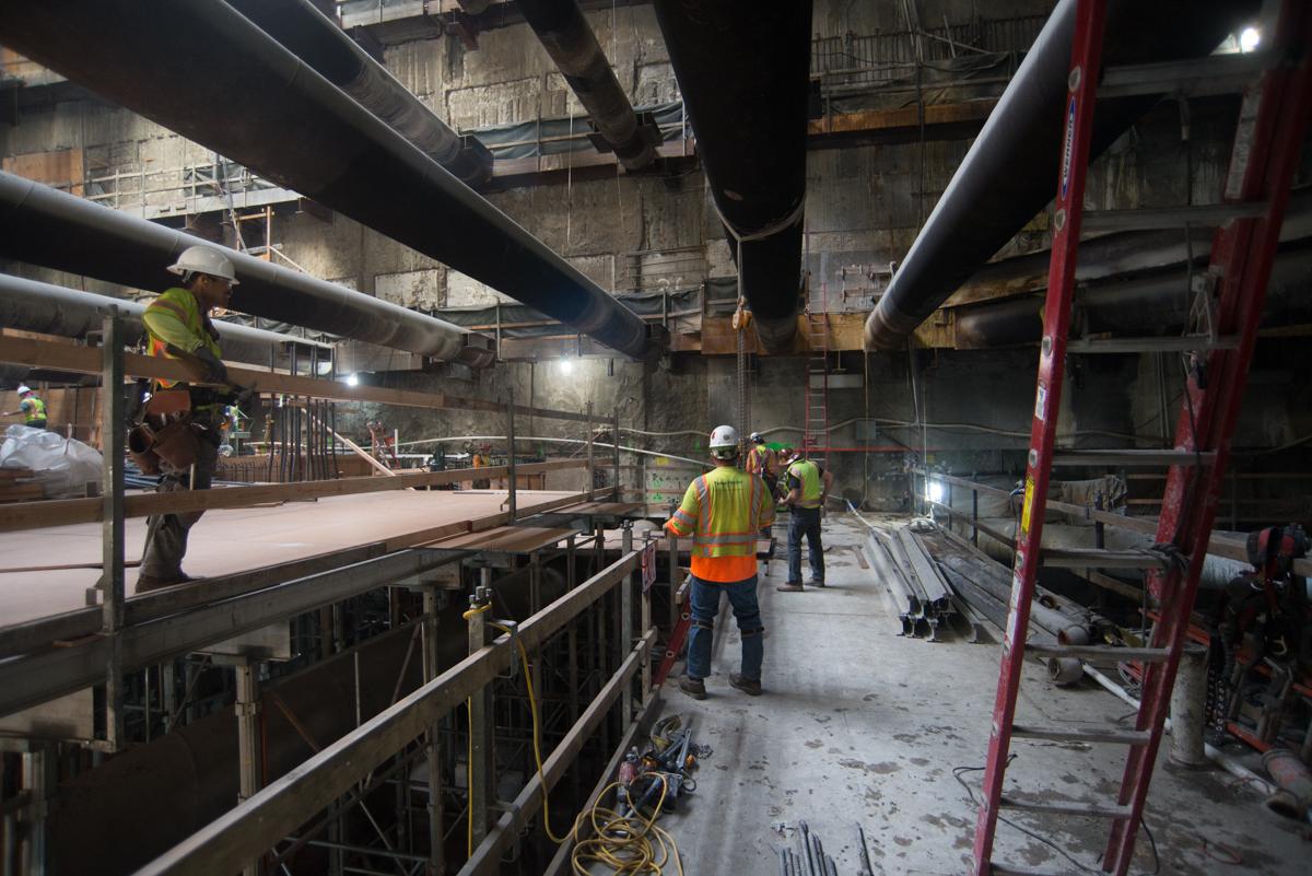 Workers in safety vests operate equipment inside an industrial station under large overhead pipes and steel structural supports.