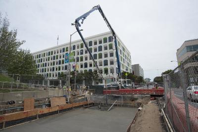Construction site with concrete pump truck, excavation equipment, and multi-story building under renovation behind red safety fencing.