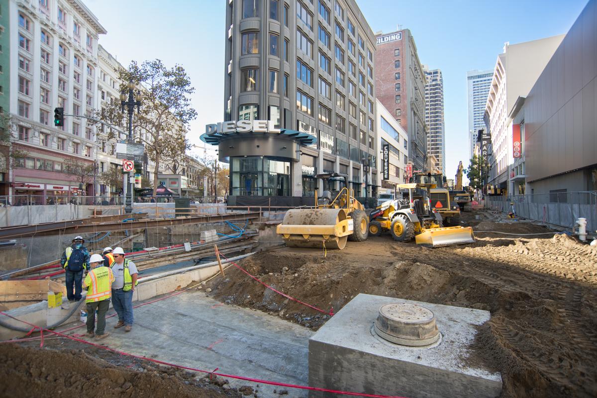 Construction site at Ellis and Stockton with workers, heavy equipment, and excavation for pile installation in downtown urban area.