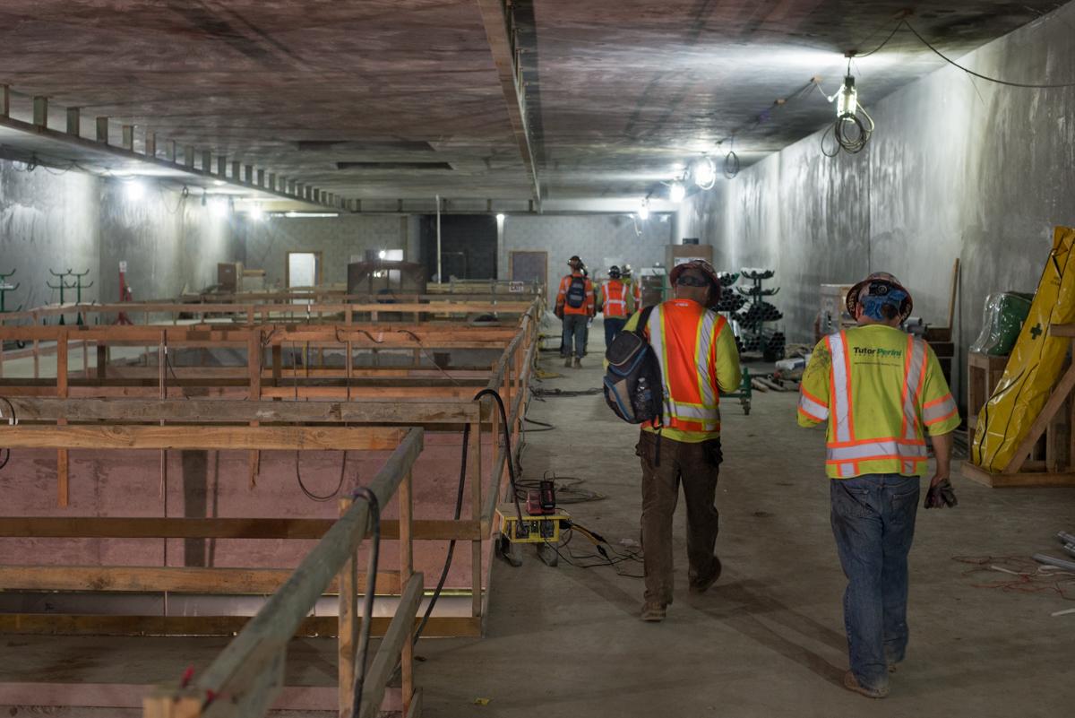 Workers in safety vests walk through an underground transit station under construction with temporary bracing and rebar structures visible.