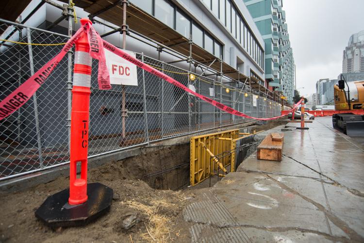 Construction site on 4th Street with red safety fencing, warning signs, and equipment along wet pavement near buildings.