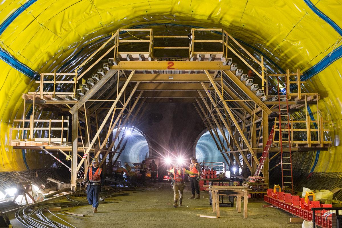 Construction workers install rebar and waterproofing inside a large yellow tunnel structure using wooden scaffolding.