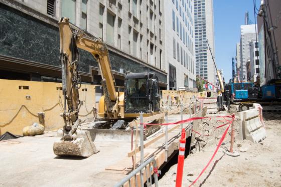 Yellow excavator on 4th Street construction site with barriers and downtown buildings in background.