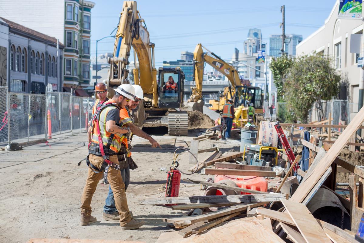 Construction workers and excavators at utility work site on 4th Street with city skyline in background.