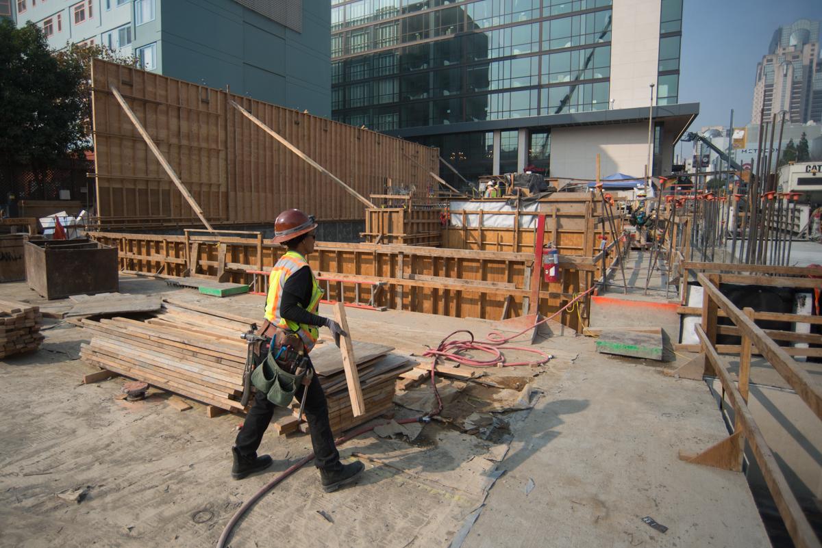 Construction worker in safety gear handling lumber on active transit station construction site with wooden formwork and metal containers.