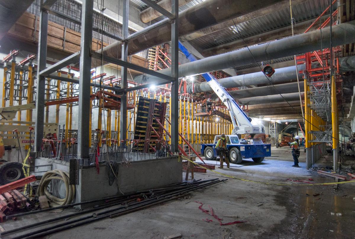 Industrial warehouse interior with steel framework, yellow safety railings, large pipes overhead, and blue mobile crane during construction work.