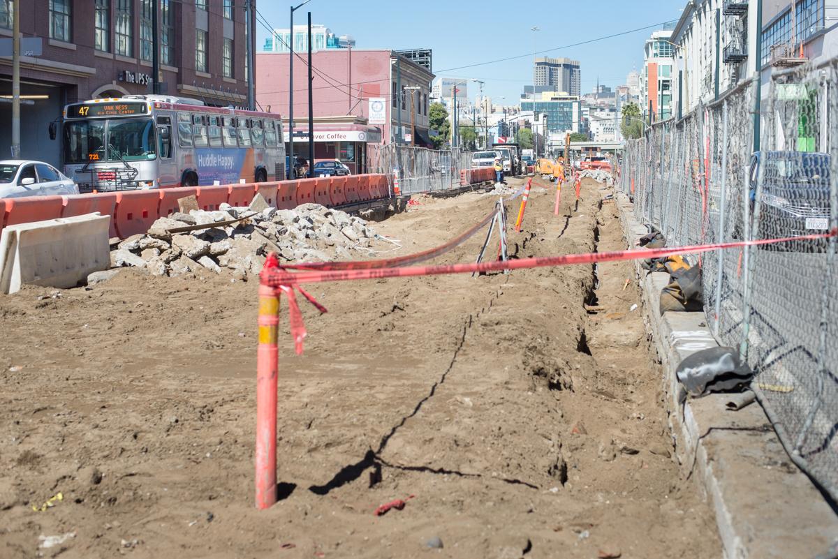 Construction site with excavated trenches, red safety barriers, and equipment along an urban street.