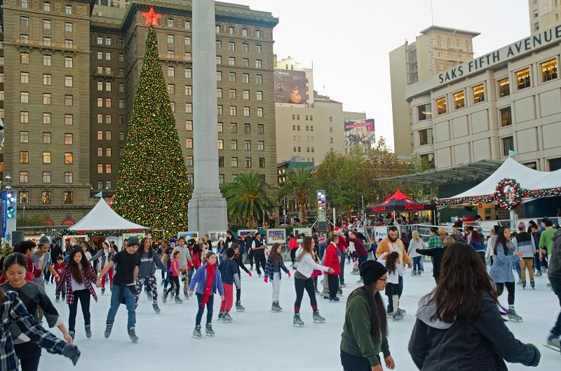 Crowds ice skating at Union Square rink with decorated Christmas tree and Saks Fifth Avenue building visible.