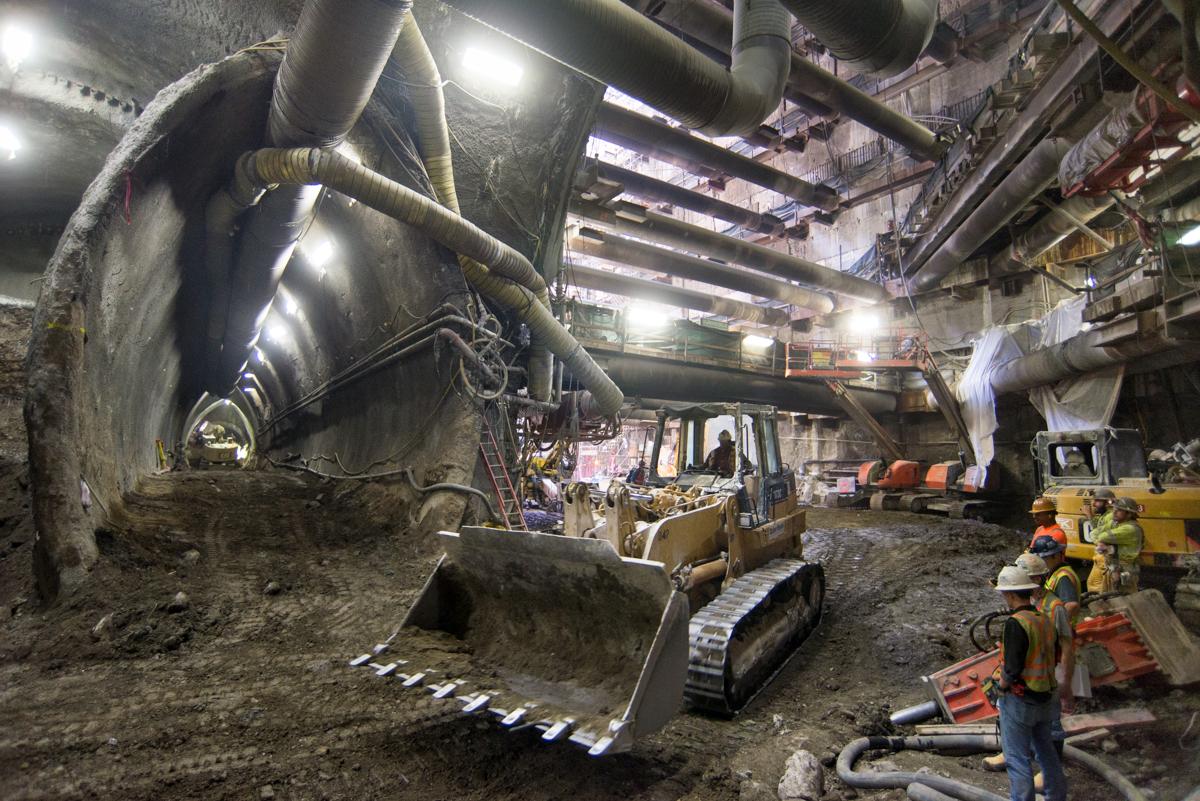 Excavator with bucket removing soil from underground transit tunnel construction site with workers and overhead lighting.
