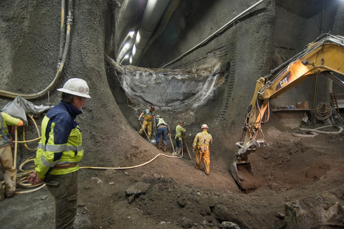 Workers in safety gear excavate underground tunnel section with yellow excavator during Central Subway construction.