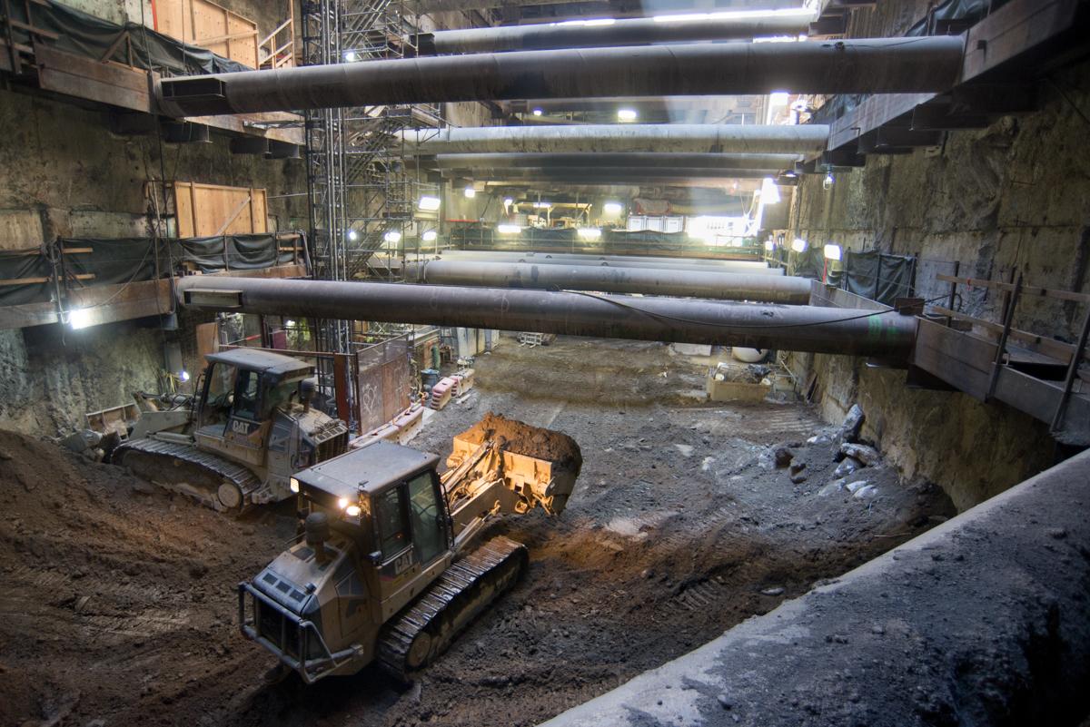 Bulldozer collecting excavated material inside underground transit station construction site with overhead pipes and lighting.