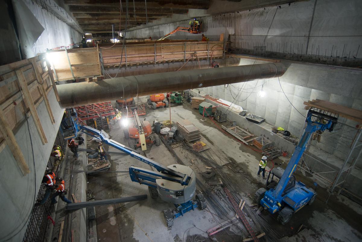 Construction workers prepare concrete forms in underground station box with large pipe and equipment visible.