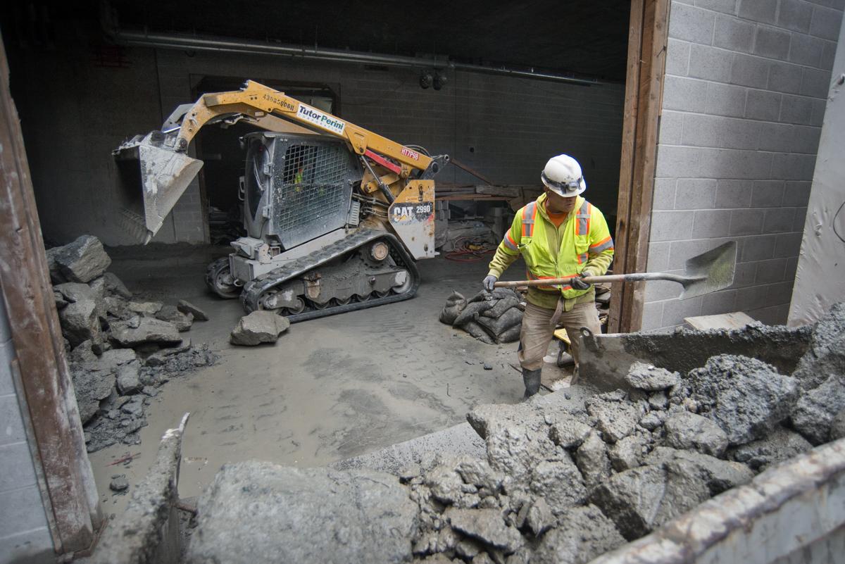 Worker with shovel and bobcat loader removing concrete debris from underground transit station mezzanine.