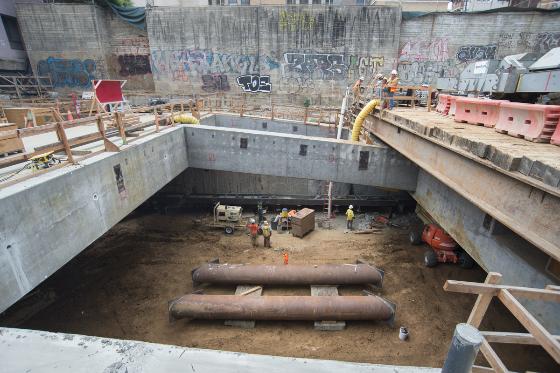 Underground excavation pit with concrete walls, wooden support beams, and construction workers at Chinatown Station site.