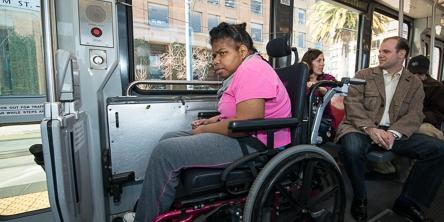 Passenger using wheelchair in designated accessible seating area on light rail vehicle with other riders nearby.