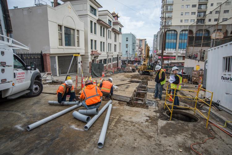 Construction workers in safety vests install large utility pipes on a closed urban street lined with buildings.