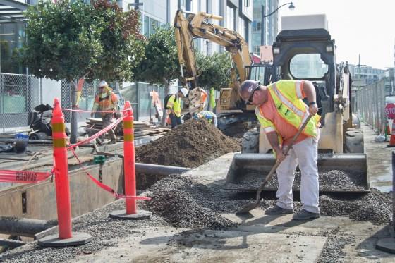 Construction workers in safety vests install sewer pipe in urban trench with excavator and equipment nearby.