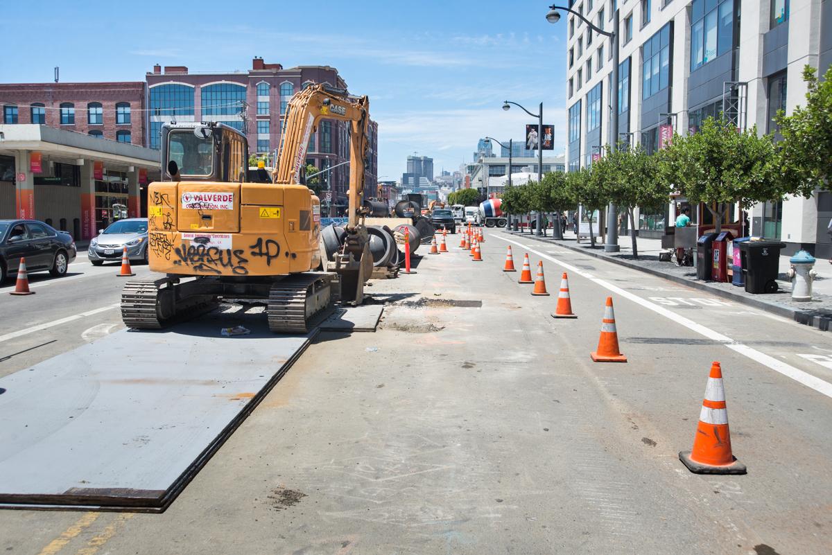 Yellow excavator on 4th Street performing sewer line installation work, surrounded by traffic cones and protective barriers.