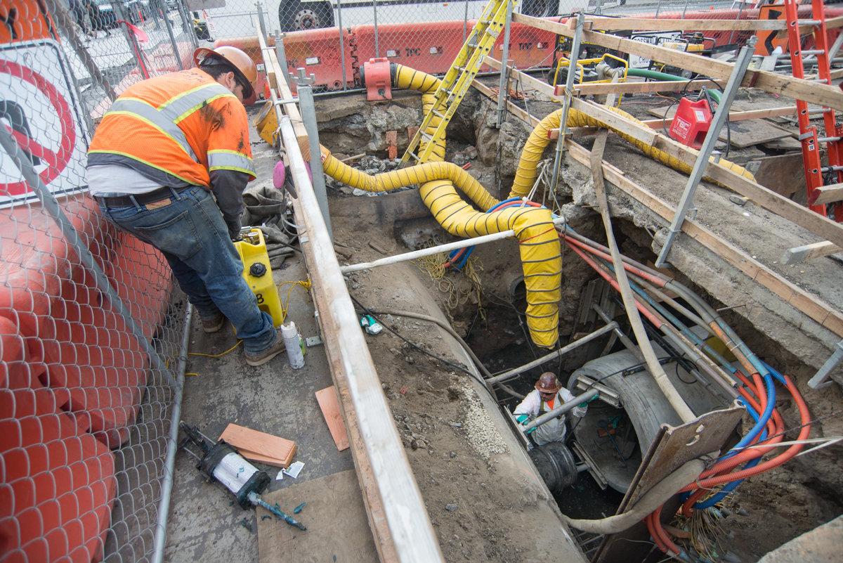 Worker in orange vest inspecting underground utility trench with yellow ventilation ducts and exposed pipes.