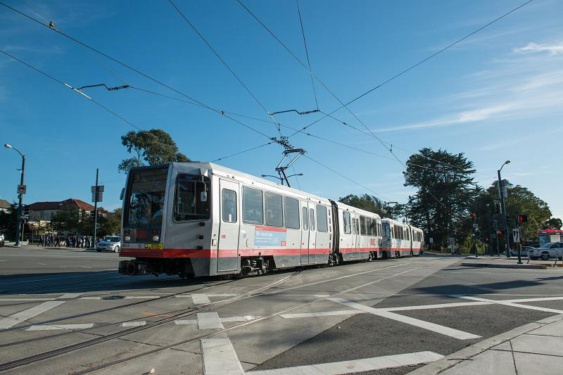 White and red Muni light rail train crossing an intersection on overhead wire tracks under clear blue sky.