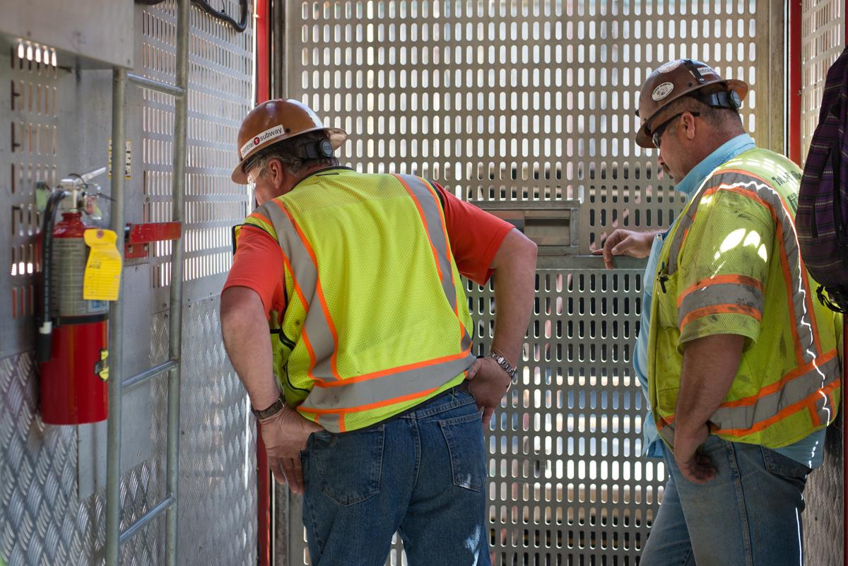 Two construction workers in hard hats and safety vests inspect equipment inside a metal cage at a transit construction site.