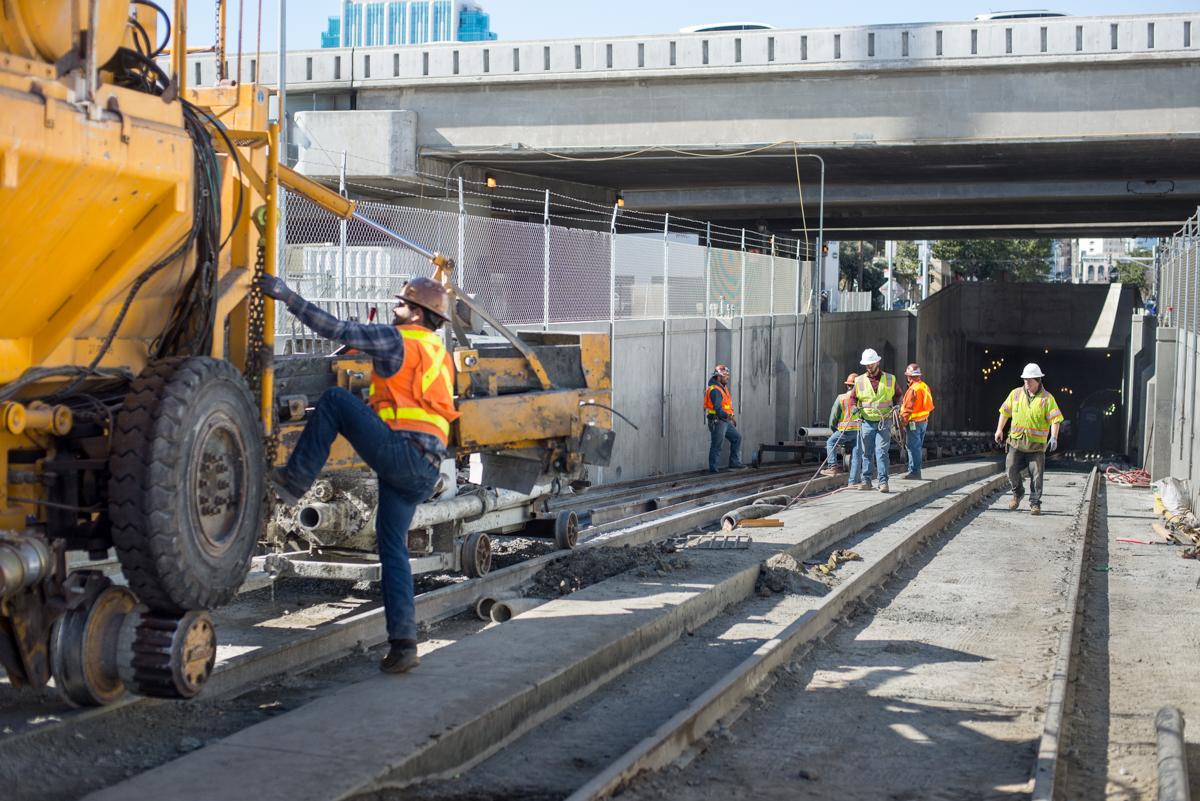 Construction workers pour concrete in transit tunnel under highway overpass.